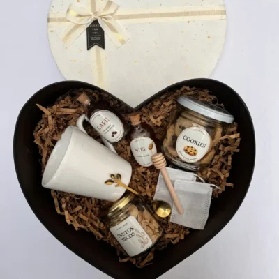 Box de regalo con forma de corazón abierto con taza blanca, café, miel, cookies y frutos secos junto a su tapa con moño.