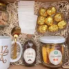 box-desayuno-dia-de-la-madre-detalle-dulzuras-cafe Detalle interior del box con botella de café molido, frasco de amarettis, frasco de cookies y bombones dorados en estuche de corazón.